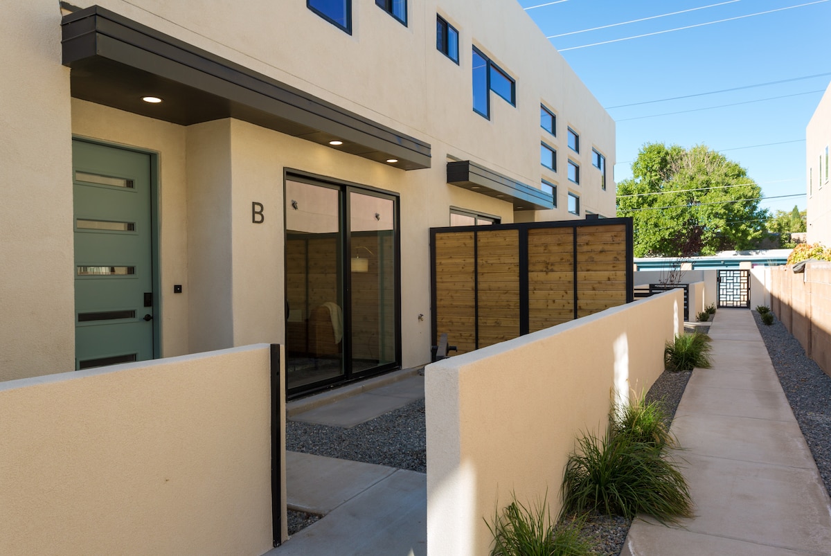 The exterior of the condo features a modern design with a frosted blue entry door. A private patio area is enclosed by wooden fencing, while a pathway lined with gravel and small plants leads to the entrance. Large windows permit natural light into the space.