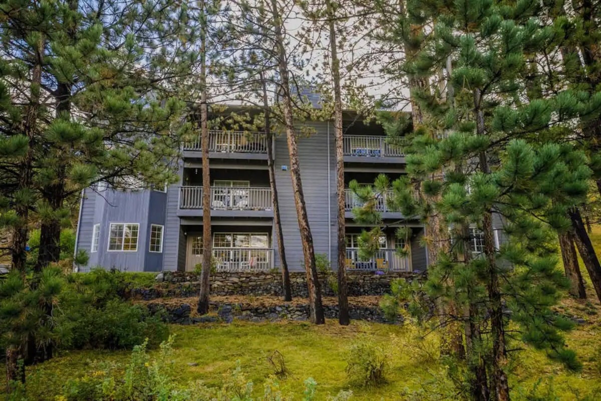 A ground-floor unit is surrounded by mature pine trees and lush greenery. The exterior features a light grey facade with multiple balconies visible on the second and third floors. Natural stone landscaping complements the serene setting.
