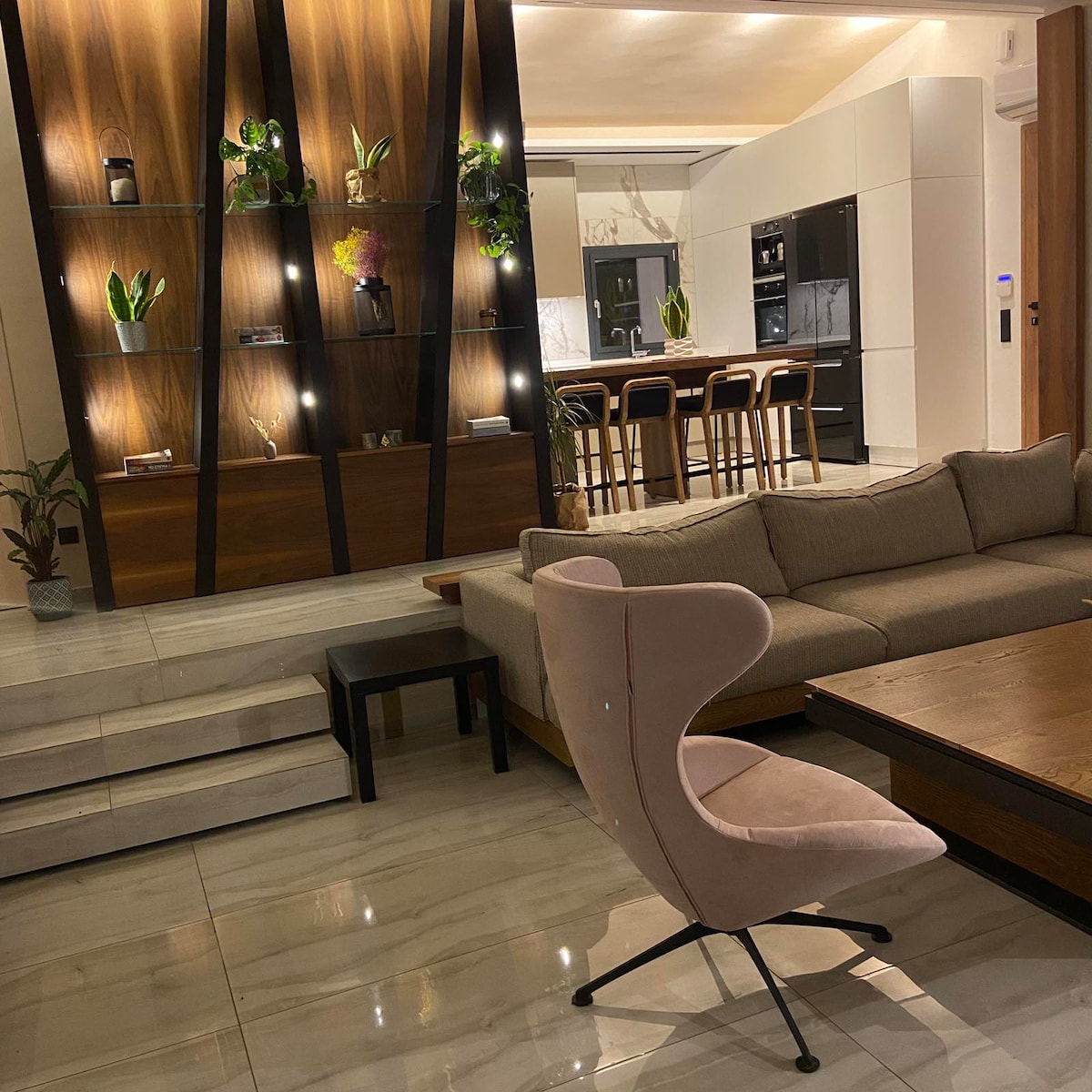 A spacious living area is highlighted by a contemporary lounge chair and a large, muted sofa. Decorative plants are displayed on a stylish wall unit, while a dining area with wooden chairs is visible in the background. Elegant marble flooring enhances the modern design.