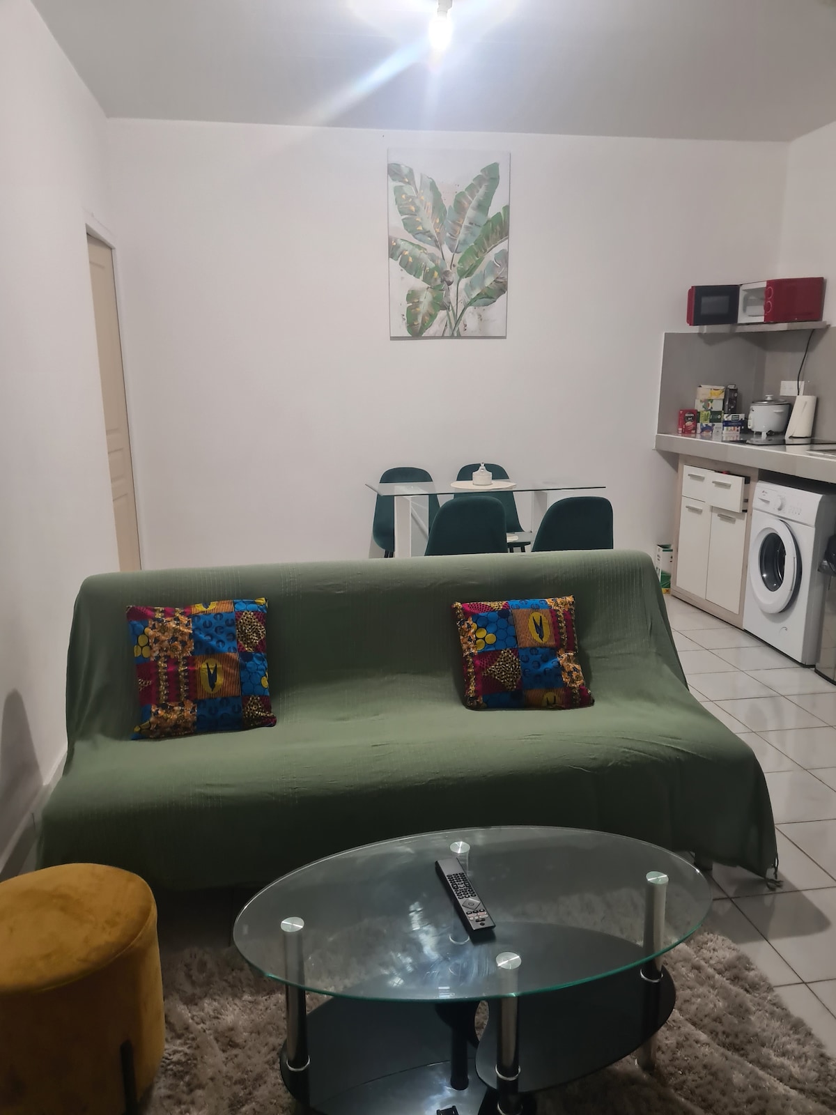 A living area is shown with a green sofa adorned with colorful cushions. A round glass coffee table sits in front, complemented by a small yellow ottoman. The space includes a dining area with a table and green chairs, with kitchen appliances visible in the background.
