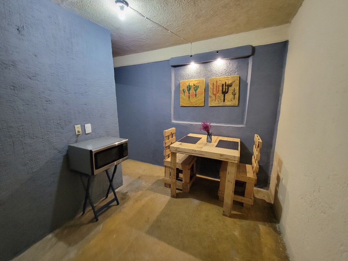 A simple dining area features a table made of wooden pallets and two matching chairs. The walls are painted in a muted blue, complemented by two cactus-themed artworks. A microwave is positioned on a stand against the wall, contributing to the functional space.