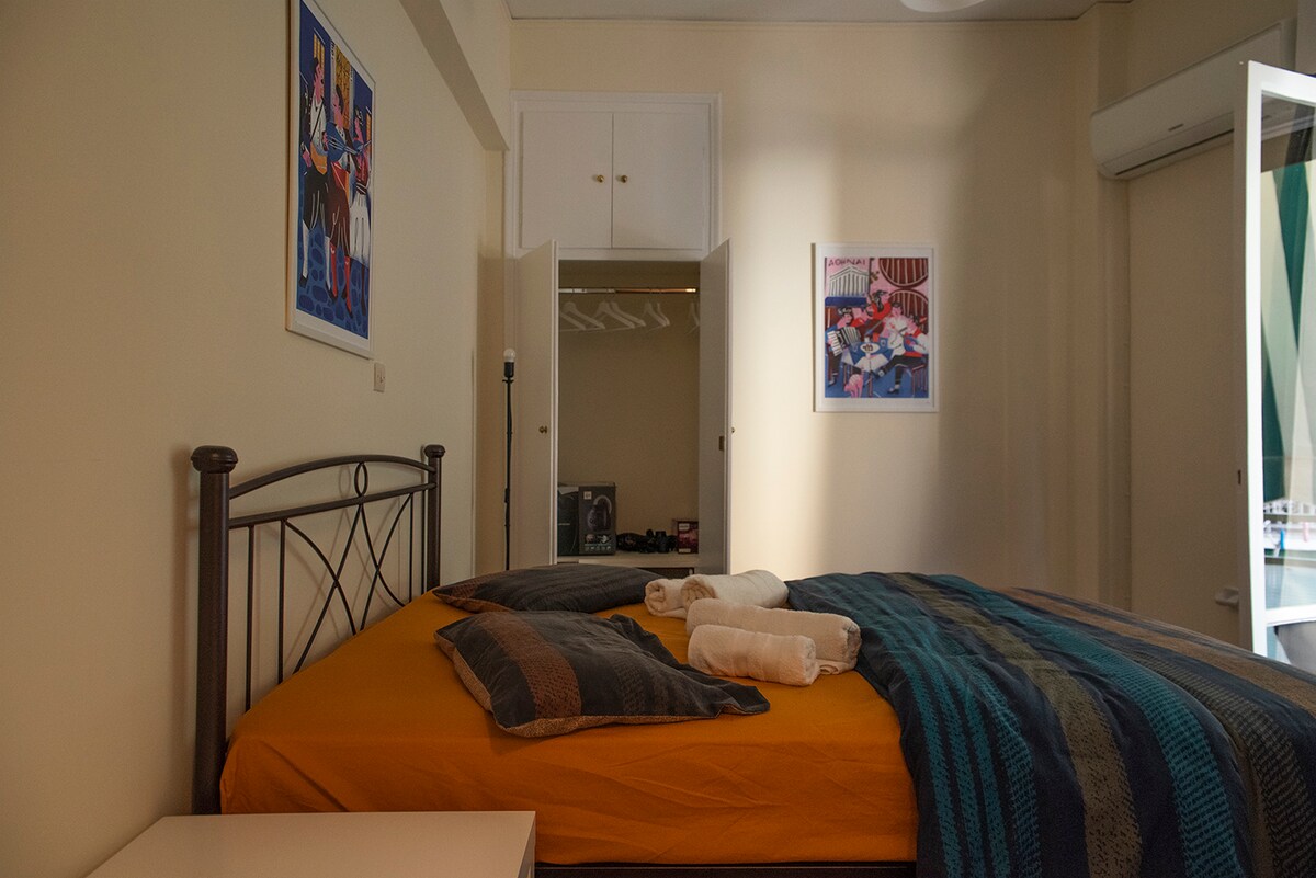 A well-lit sleeping area features a spacious bed with yellow bedding and decorative pillows. Freshly rolled towels are placed on top. In the background, a wardrobe can be seen with doors slightly ajar, revealing clothing storage. Colorful artwork adorns the walls, adding character to the room.