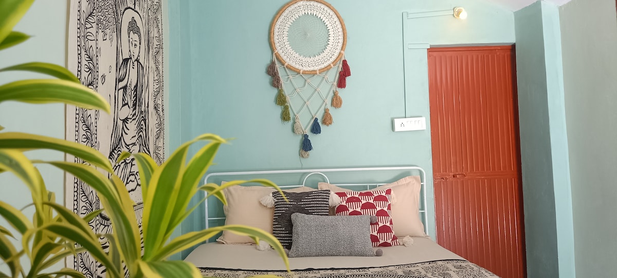 A cozy sleeping area is adorned with a large wall tapestry and a decorative dreamcatcher above the bed. A light green wall provides a calming backdrop for the neatly arranged cushions and throws on the bed, complemented by a plant situated in the foreground.