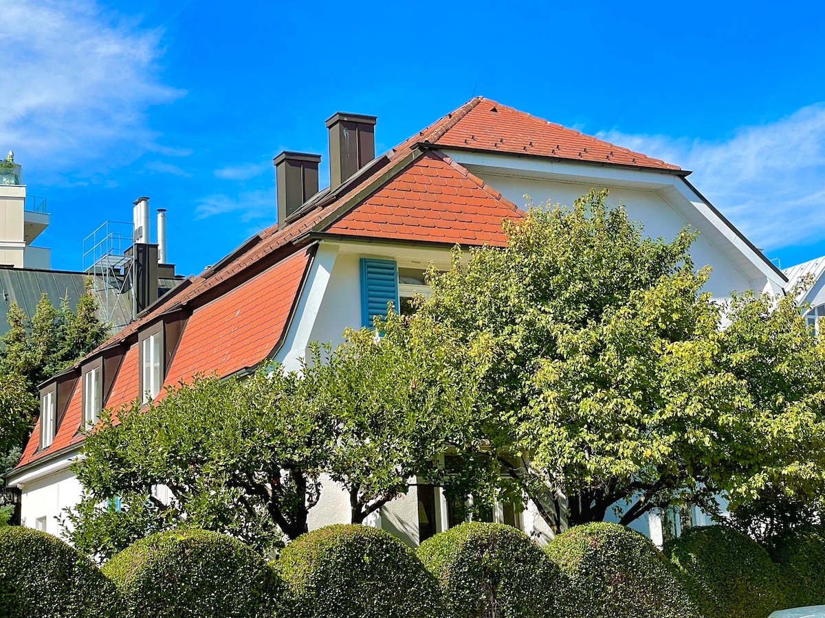 A well-maintained villa features a sloping red roof and large windows framed by lush greenery. The white exterior complements the vibrant surroundings, while neatly trimmed hedges add to the inviting atmosphere. A clear blue sky expands gracefully above the structure.