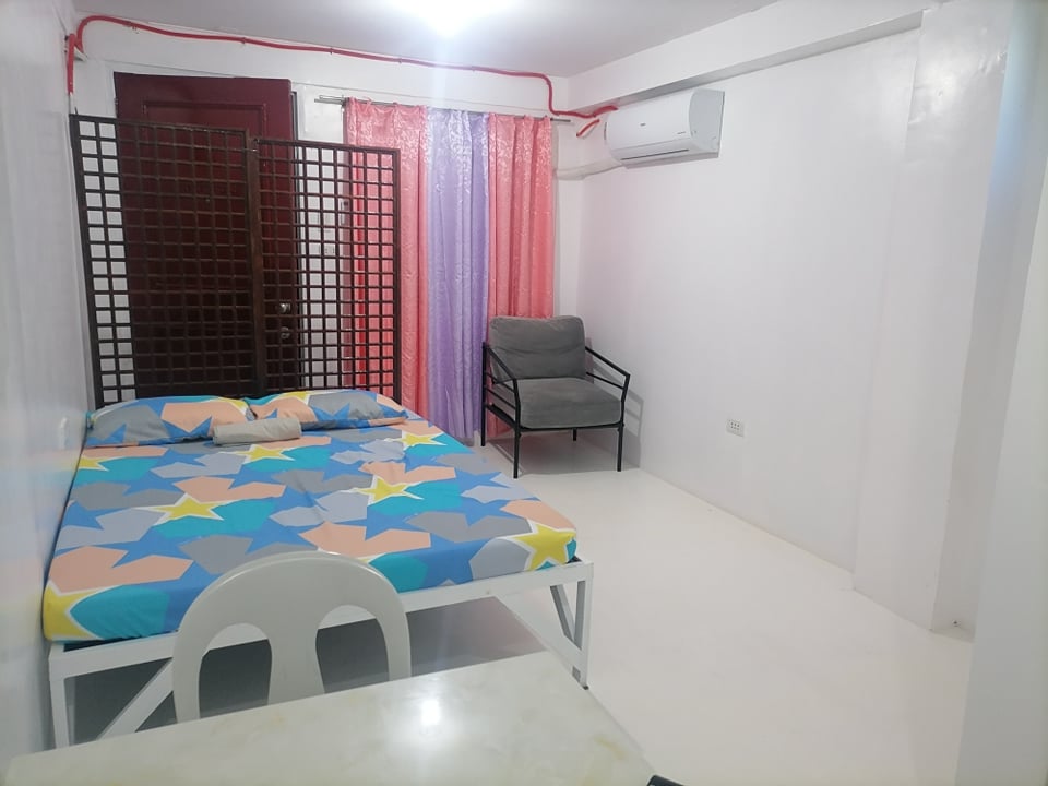 A bright room features a spacious bed with a colorful star-patterned comforter. A gray chair is positioned beside the bed. Sheer curtains in varying shades of pink hang over a window, while a small table is visible in the foreground.