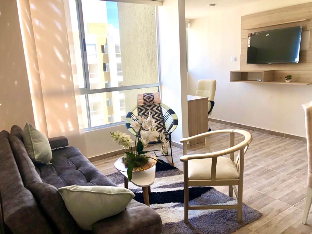 A modern living area is highlighted by a comfortable gray sofa and a round coffee table. A TV mounted on a wooden panel is visible, and natural light is streamed through large windows. A chair and a rug add texture to the space.