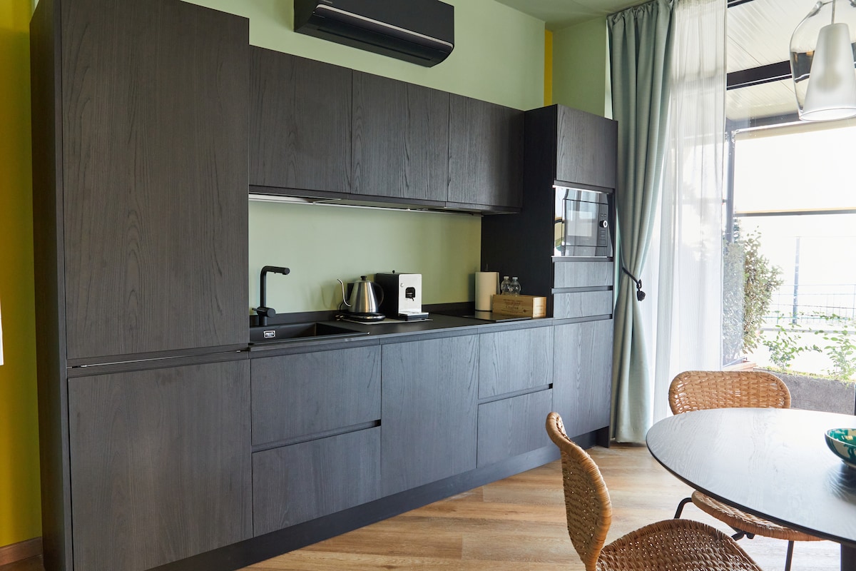 A modern kitchen area is shown with dark cabinetry and sleek countertops. Kitchen appliances are neatly arranged, including a kettle and various glass containers. Natural light enters through large windows, highlighting the light wood flooring and the dining table set with woven chairs.