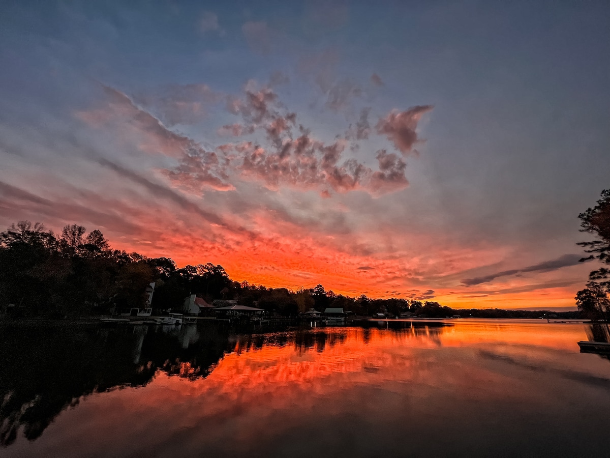 A vibrant sunset casts hues of orange, pink, and purple across the sky, reflected on the calm surface of the lake. Silhouettes of trees and structures can be seen along the shore, creating a serene atmosphere.