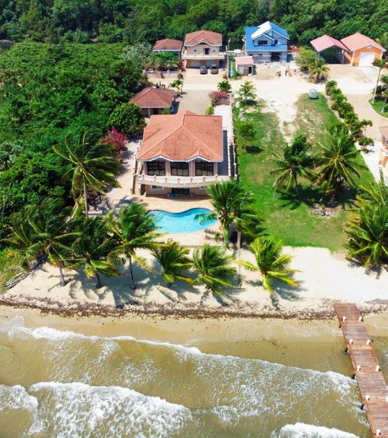 A panoramic view of the beachfront property reveals a spacious house with a red-tiled roof, surrounded by palm trees. A swimming pool in the yard is visible, along with sandy beach for direct ocean access. Adjacent houses and a pier can be seen in the distance.