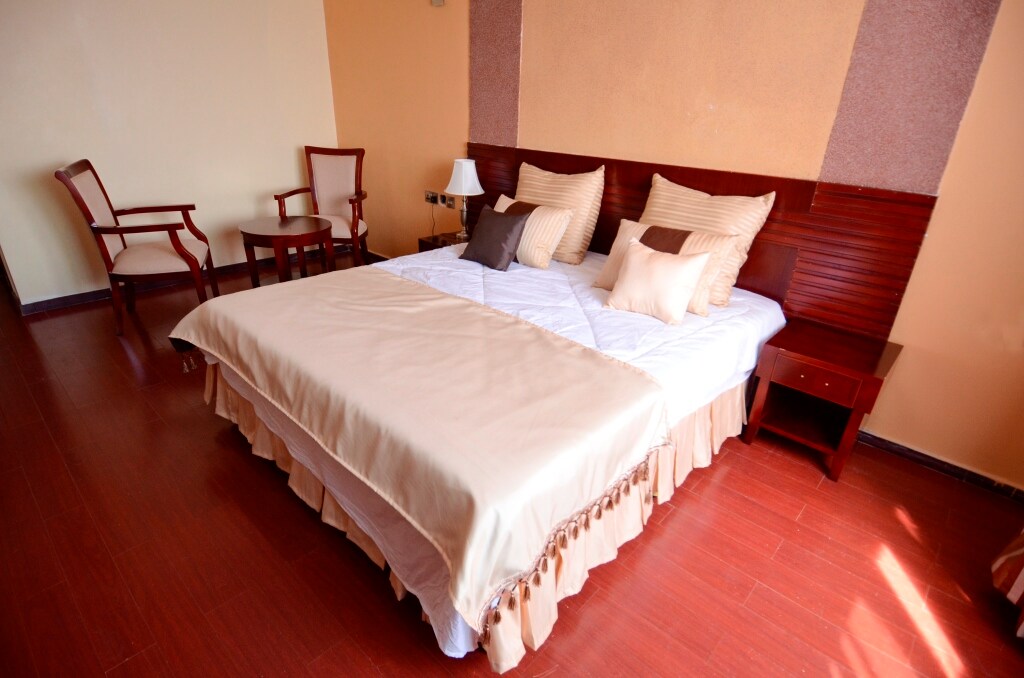 A spacious sleeping area features a bed dressed in neutral-toned linens with decorative pillows. Two wooden chairs are positioned beside a small table, adding to the inviting layout. Natural light filters in, highlighting the warm wood flooring and subtle wall textures.