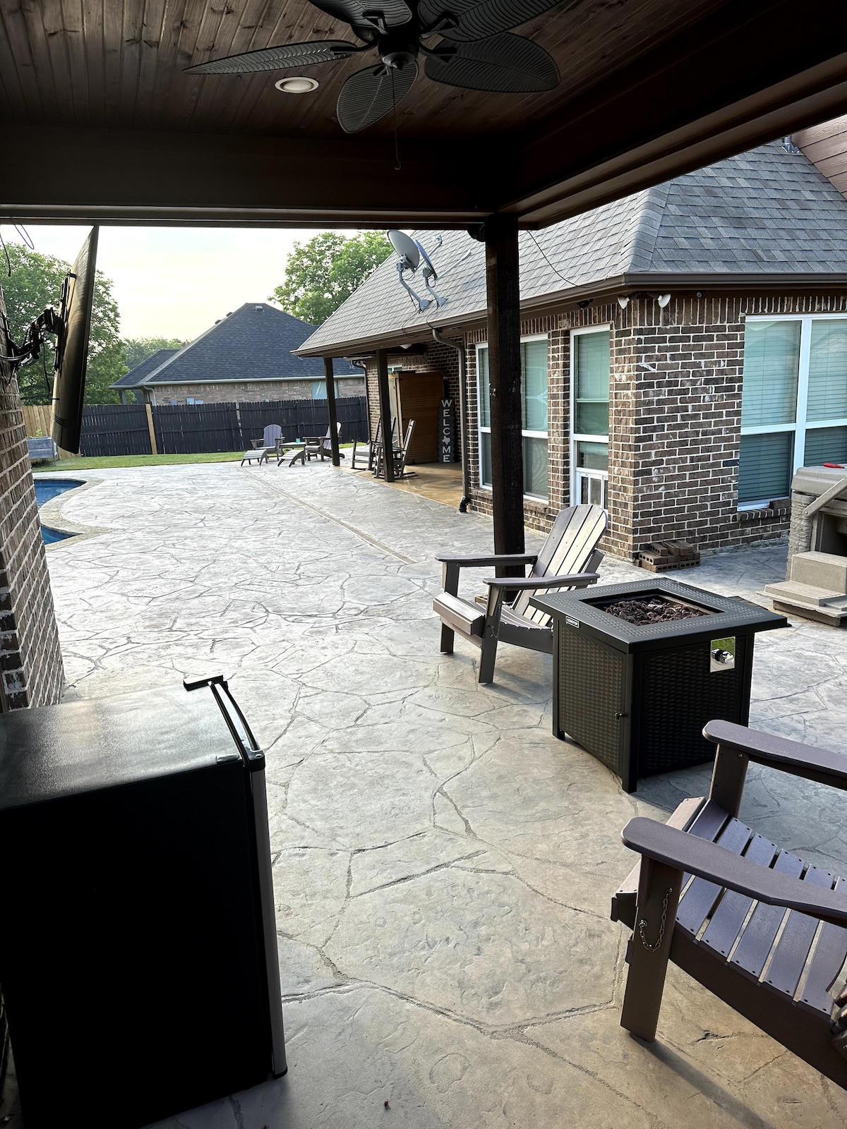 An outdoor patio area features a stone floor and wooden furniture. Two chairs sit adjacent to a fire table, and a small refrigerator is positioned against one wall. The pool area and house are visible in the background, with trees surrounding the property.
