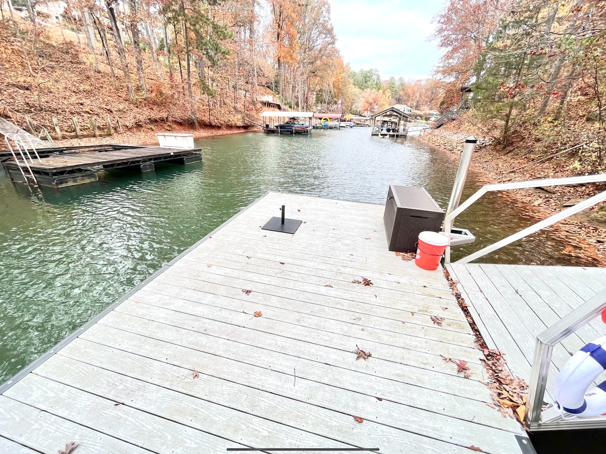 A wooden dock extends over the tranquil waters of a lake, surrounded by trees displaying fall foliage. Various boats are anchored along the shore, with a clear pathway leading into the water. A storage box and a red bucket are visible on the dock.