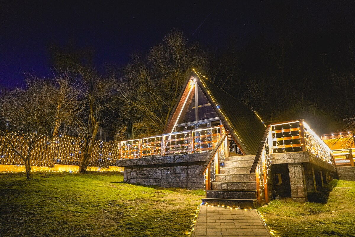 The A-frame cabin is highlighted by warm outdoor lighting, creating a welcoming atmosphere. Steps lead to the entrance, surrounded by a grassy area and trees. A deck extends from the cabin, providing additional outdoor space for relaxation.