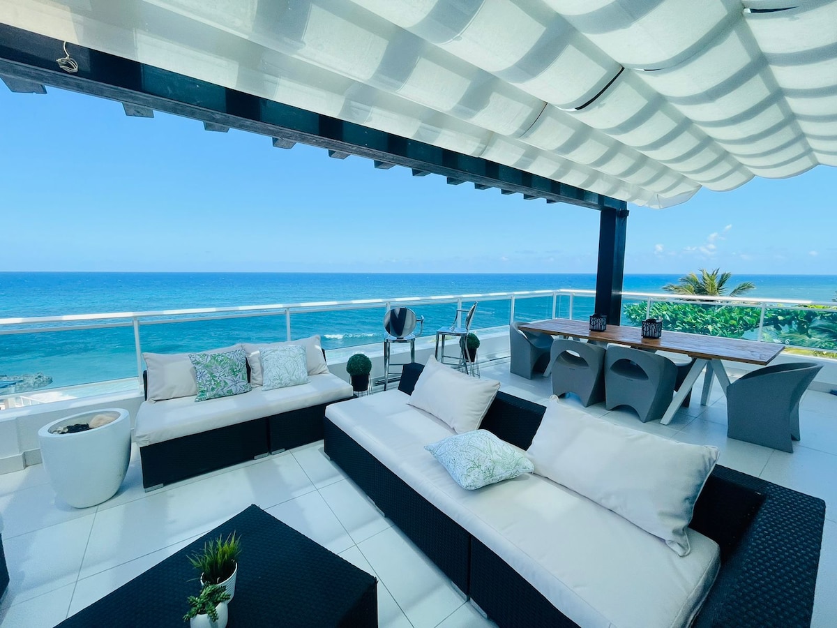 An outdoor terrace is presented with comfortable seating arrangements, featuring light-colored cushions and dark wicker furniture. A dining area is positioned next to the seating, surrounded by stunning ocean views. The overhead canopy offers shade, creating a pleasant atmosphere for relaxation.