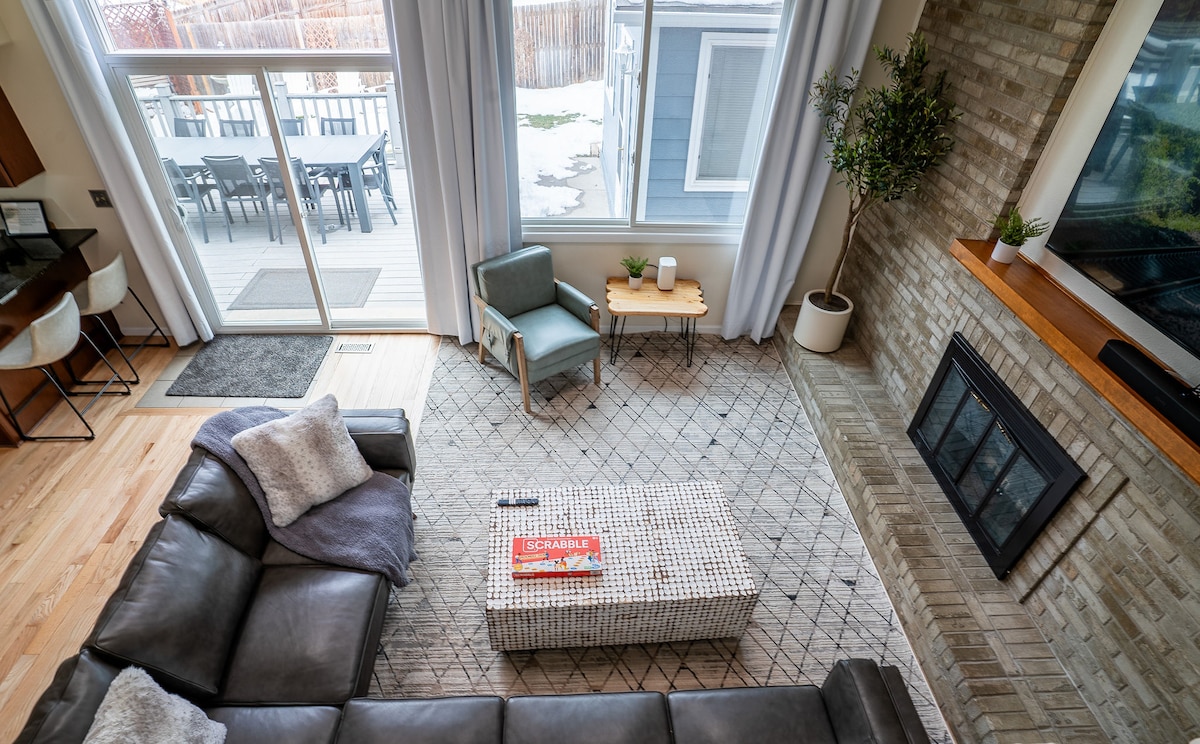 An open-concept living area is seen from above, featuring a sectional sofa, a patterned area rug, and a coffee table with games. Large windows provide natural light, while a fireplace is integrated into a brick wall. A comfortable chair sits beside a small side table.