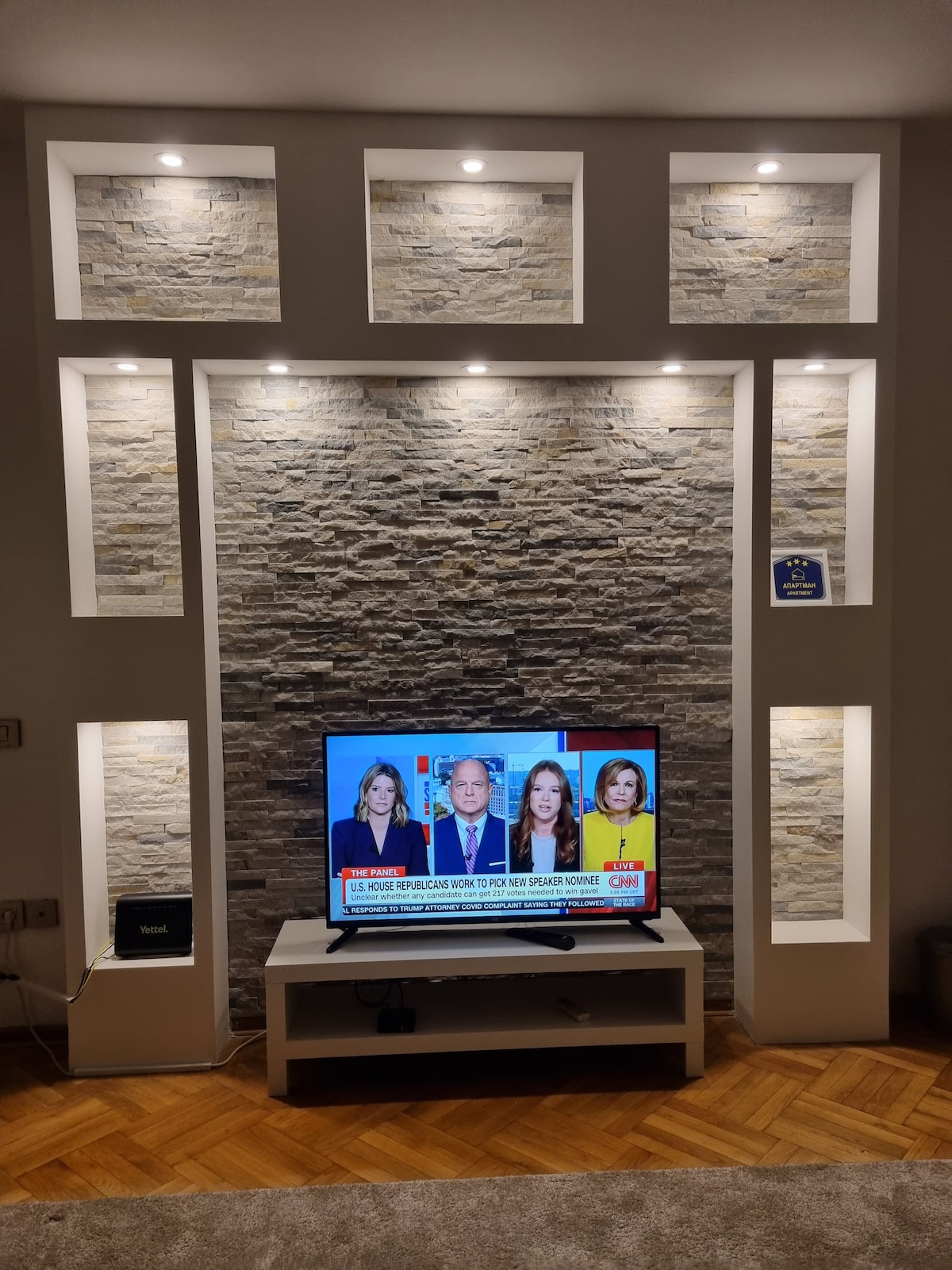 A modern entertainment area displays a flat-screen TV on a minimalist media unit. The wall features stone accents and illuminated shelves that provide ambient lighting. A plush area rug lays beneath, contributing to the cozy setting.