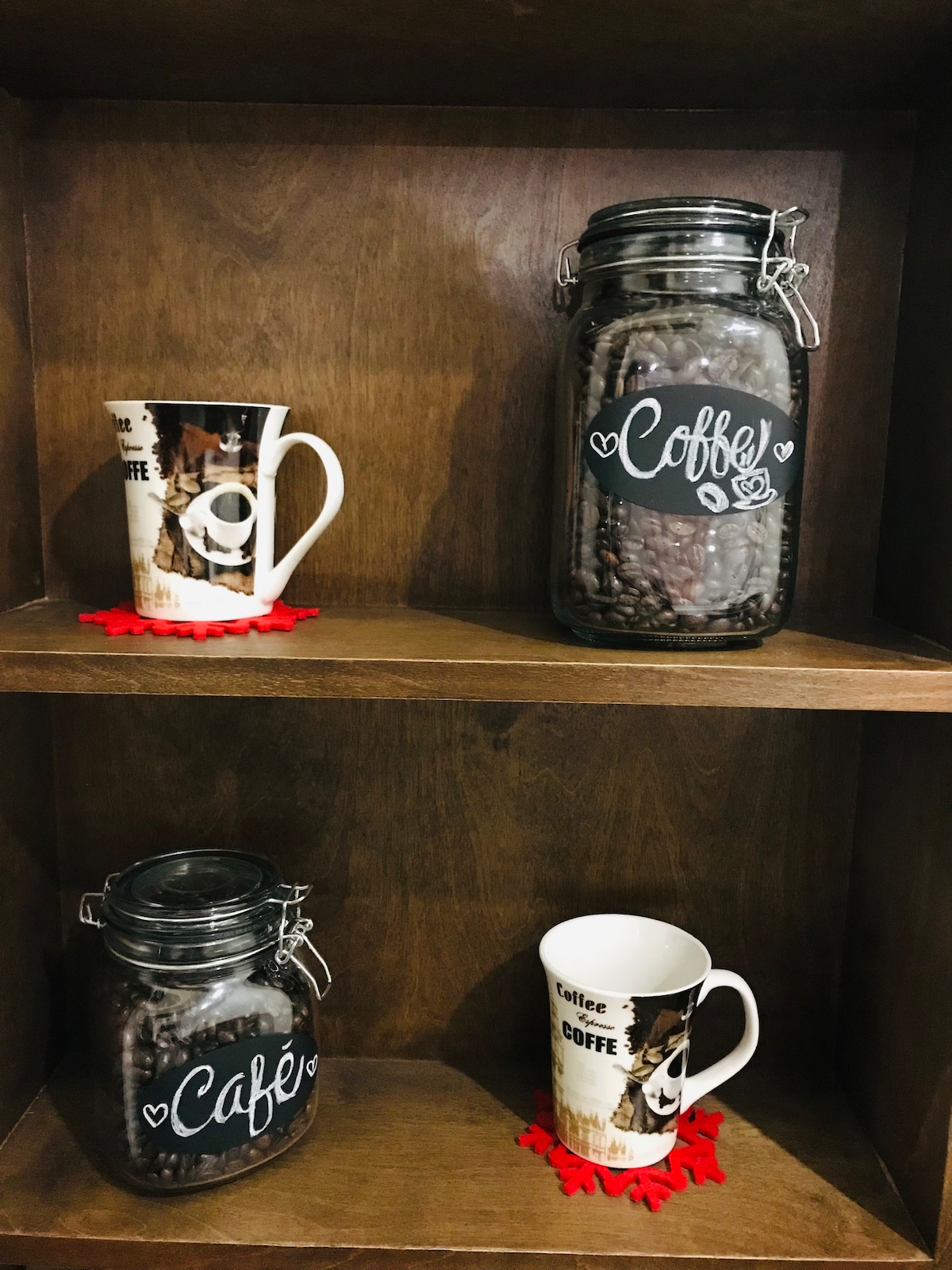 A wooden shelf displays two glass containers filled with coffee beans, one prominently labeled 'Coffee' and the other 'Café.' Adjacent, a large coffee mug features a playful coffee-themed design. The items are set against a warm-toned backdrop, enhancing their visibility.