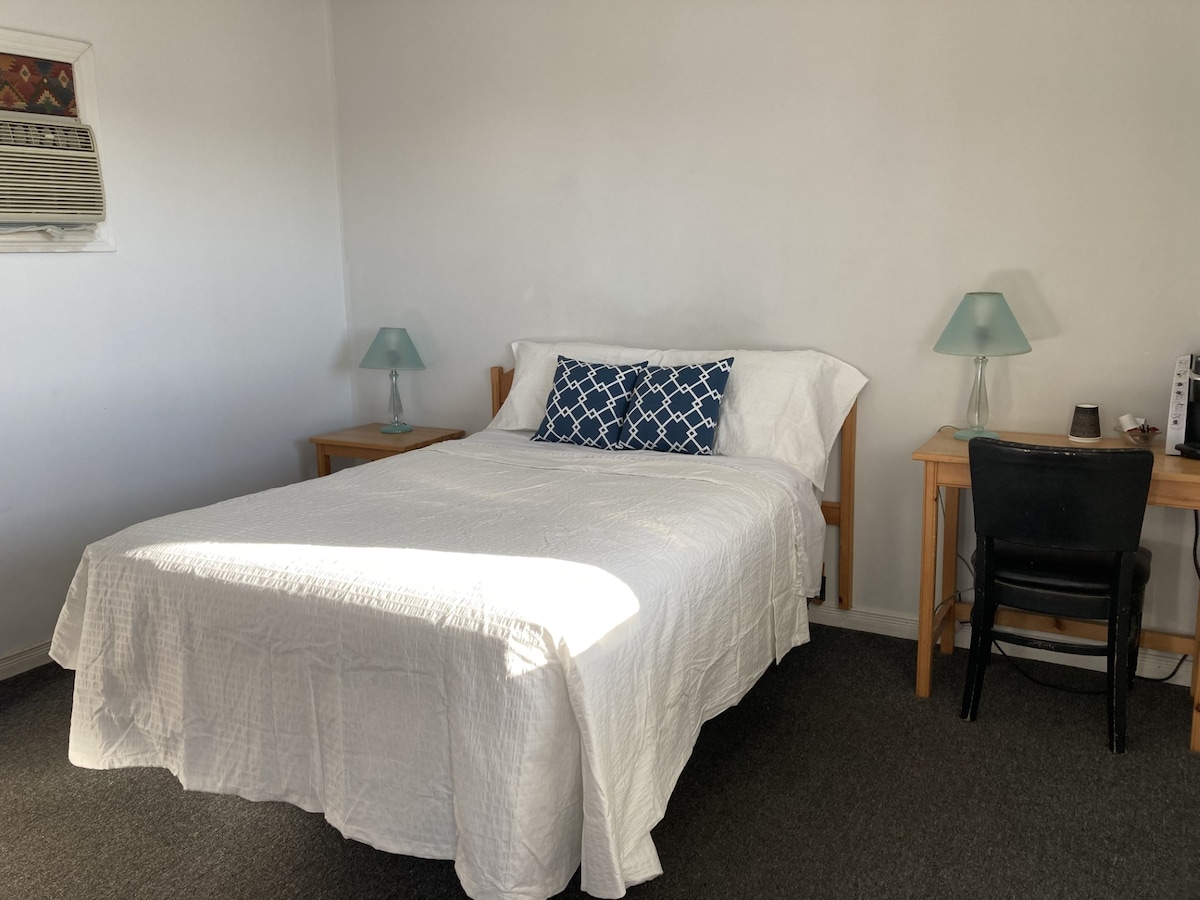 A simple room features a double bed with a white quilt spread and a decorative blue pillow. A small desk and chair provide workspace, while two bedside tables are positioned on either side of the bed. Natural light enters through a window, enhancing the neutral tones of the space.