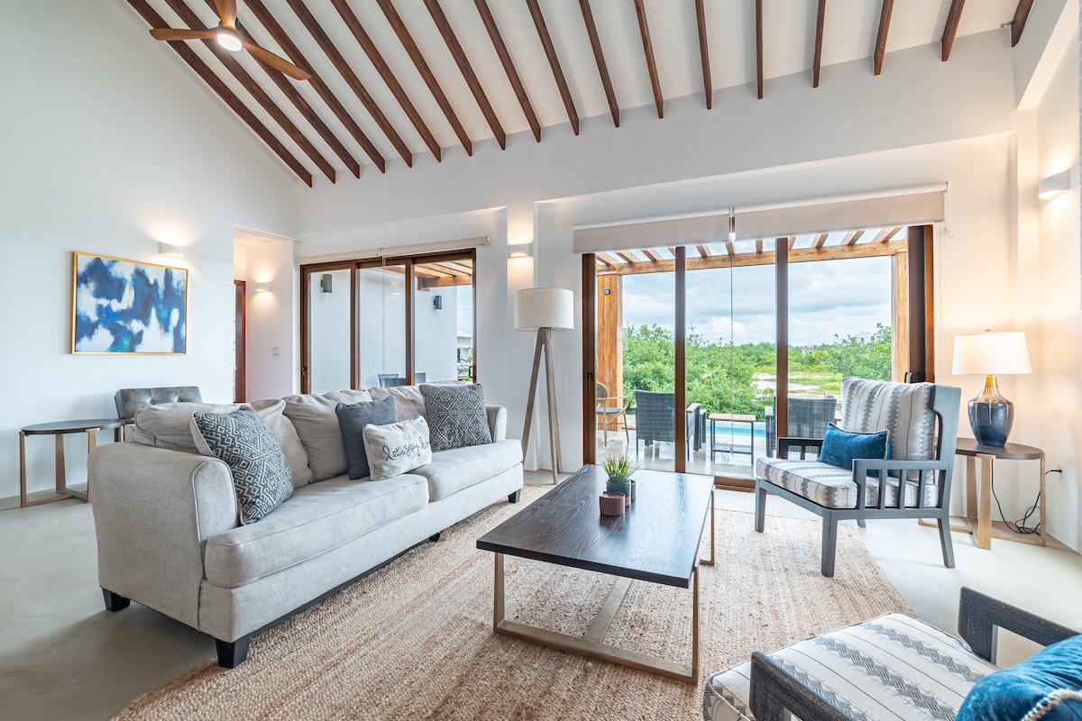 A spacious living area features high wooden beams and large glass doors that open to an outdoor view. A light-colored sofa is adorned with decorative pillows, while a wooden coffee table is positioned in the center. Natural light fills the room, enhancing the minimalist decor.