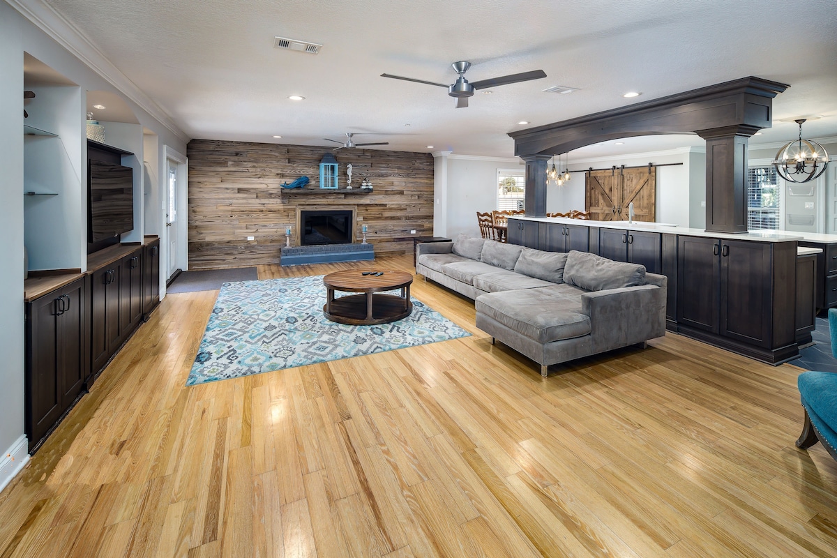 A spacious living area features hardwood floors and a warm wooden accent wall. A large grey sectional sofa is positioned in front of a modern coffee table, with a fireplace serving as a focal point. Light fixtures and open shelving contribute to the inviting atmosphere.
