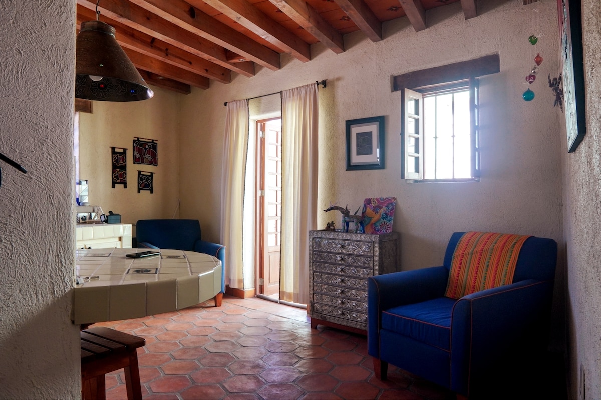 A living area features blue seating options, including a sofa and an armchair. Natural light enters through two windows and a door, illuminating the textured walls and terracotta tile flooring. Decorative wall art and a wooden ceiling add character to the space.