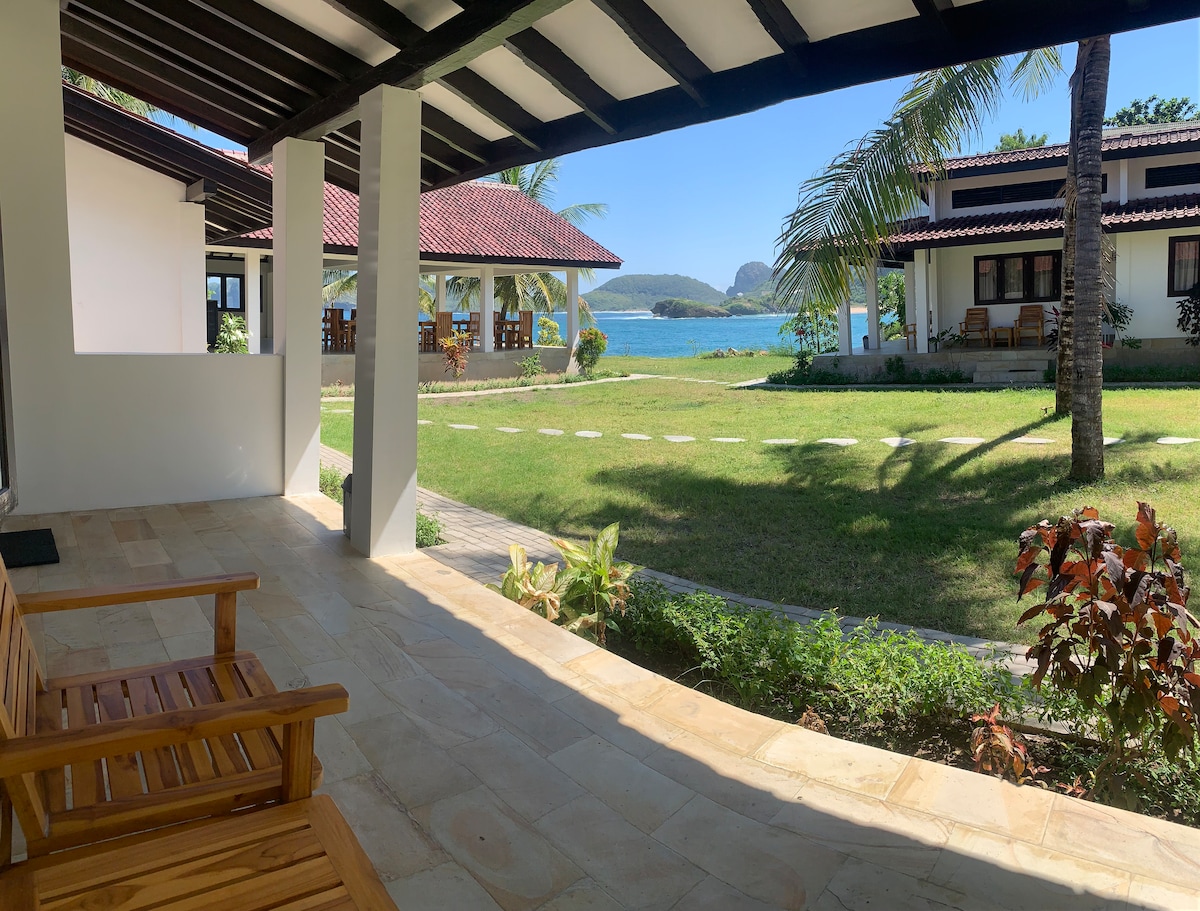 A covered patio area showcases two wooden chairs, offering views of a lush green lawn and the water beyond. Palm trees frame the scene, with additional buildings visible in the background, highlighting the serene coastal environment.