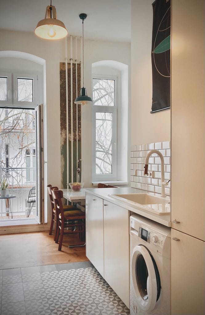 A modern kitchen space is visible, featuring sleek cabinetry and a contemporary sink. A washing machine is integrated into the cabinetry. Natural light enters through large windows, illuminating a small dining table with wooden chairs near the balcony. Decorative tiles add texture to the floor.