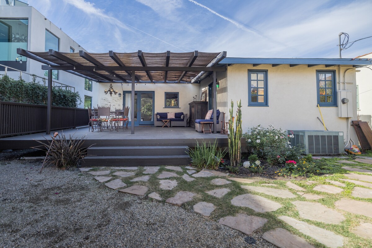 A spacious outdoor deck is highlighted, featuring shaded seating areas under a pergola. A dining table is set among greenery and colorful flowers, with a stone pathway leading through the yard. The building showcases a light-colored exterior and multiple blue-framed windows.
