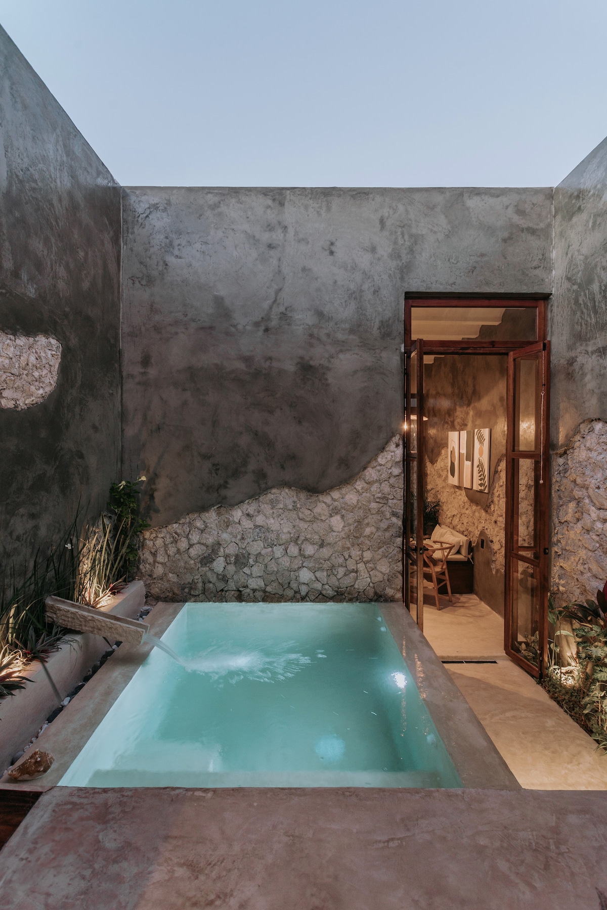 A serene private pool is framed by smooth, textured walls, reflecting a calming blue hue. An inviting doorway leads to a cozy dining area. Soft lighting enhances the natural elements, creating a tranquil outdoor space that encourages relaxation.