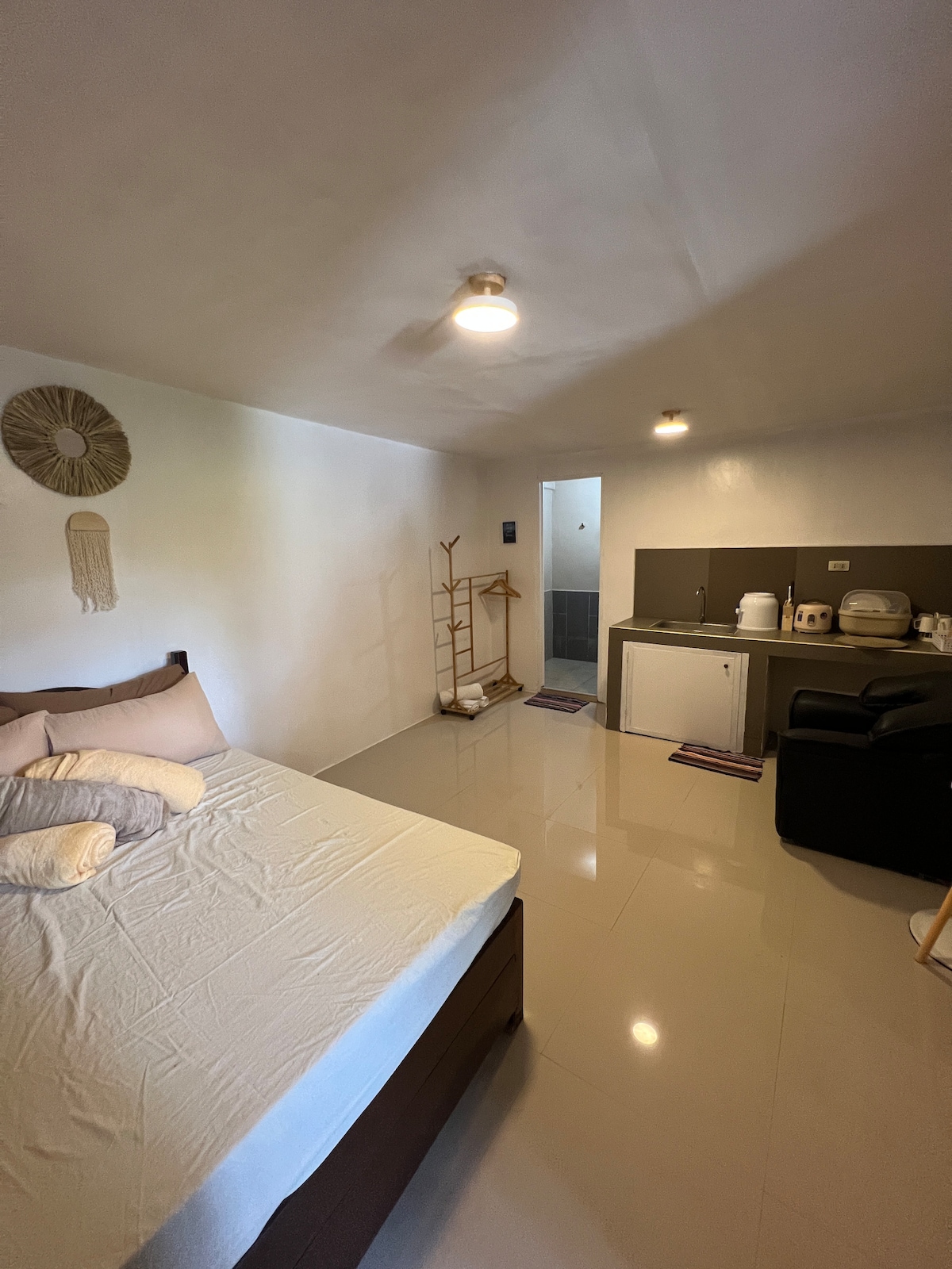 A newly renovated studio unit features a spacious bed with neutral bedding. A compact kitchenette is visible, equipped with essential utensils. Soft lighting enhances the ambiance, while a doorway leads to a private bathroom. A minimalist clothing rack is positioned beside the bed.