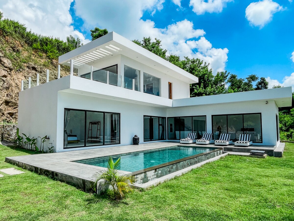 The two-story villa features a modern architectural design with large glass windows allowing natural light. An expansive infinity pool lies adjacent to the building, surrounded by a well-maintained lawn and tropical plants. Comfortable sun loungers are positioned by the pool, offering an enjoyable outdoor space.