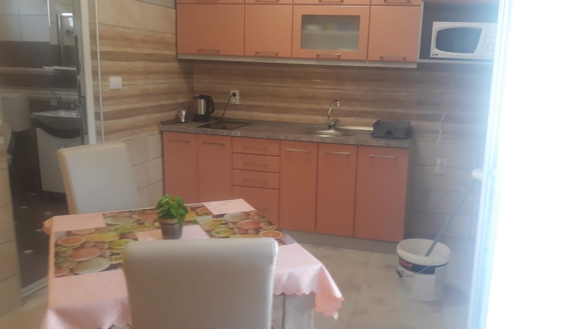A modern kitchen features orange cabinetry and a marble countertop. A small dining table with a floral tablecloth is accompanied by two white chairs. Essential appliances, including a microwave and kettle, are available. Natural light enters through a nearby door.
