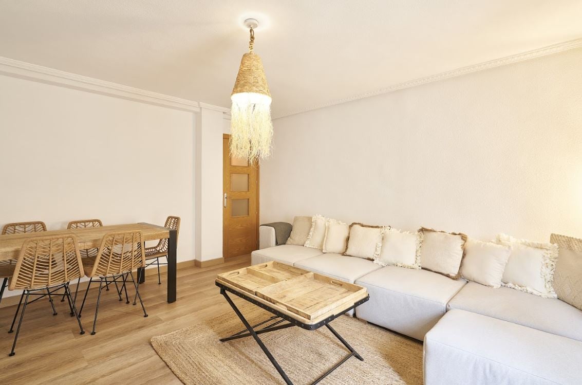 A spacious living area features a light-toned sectional sofa with soft cushions and a wooden coffee table in the center. A warm pendant light hangs from the ceiling, while a dining table with woven chairs is visible in the background, enhancing the room's welcoming ambiance.