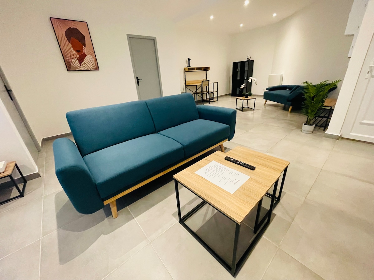 A spacious living area is shown, featuring a teal sofa and a wooden coffee table with a remote control on it. Modern decor elements are visible, including a framed artwork on the wall and additional seating in the background.