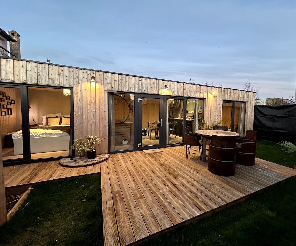 A wooden house exterior is shown, featuring large glass doors that provide access to a cozy interior. A spacious wooden deck surrounds a circular dining table, complemented by chairs. Soft lighting illuminates the exterior, enhancing the inviting atmosphere during twilight.