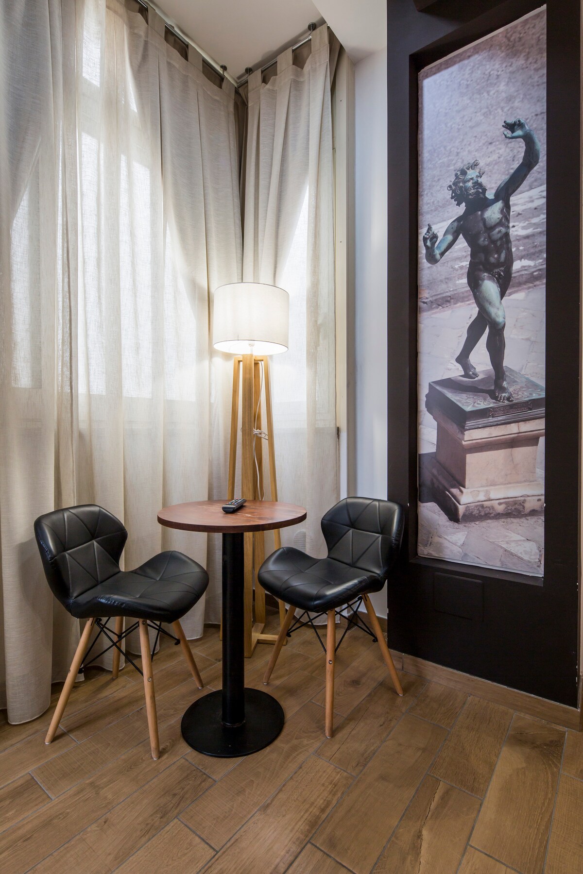 A small corner is highlighted by a round wooden table surrounded by two black chairs with a modern design. A tall lamp provides soft illumination, while sheer curtains allow natural light to filter through. A wall mural features a classical statue, adding cultural interest.