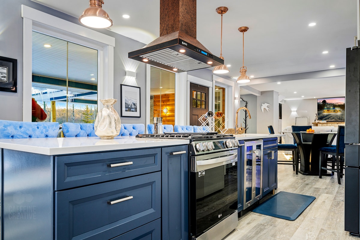 A modern gourmet kitchen is highlighted by a large island with seating and a sleek stovetop hood. Stainless steel appliances, including a professional-grade range, complement the blue cabinetry. Natural light floods the space through wide windows, creating an inviting atmosphere for cooking and gathering.