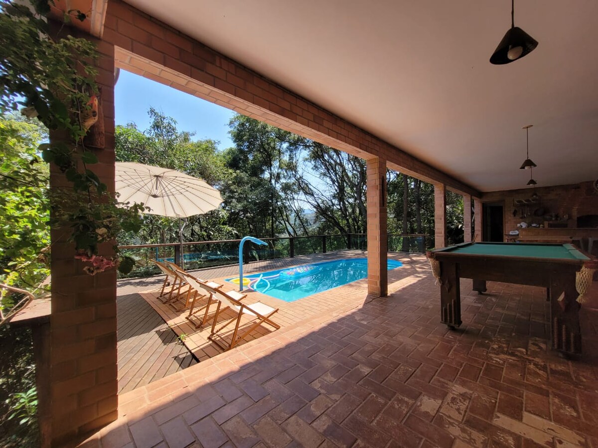 A shaded outdoor area features a turquoise pool surrounded by lush greenery. Wooden lounge chairs are positioned beneath a large umbrella, while a game table is seen nearby. Bright sunlight filters through the open space, enhancing the relaxed atmosphere.