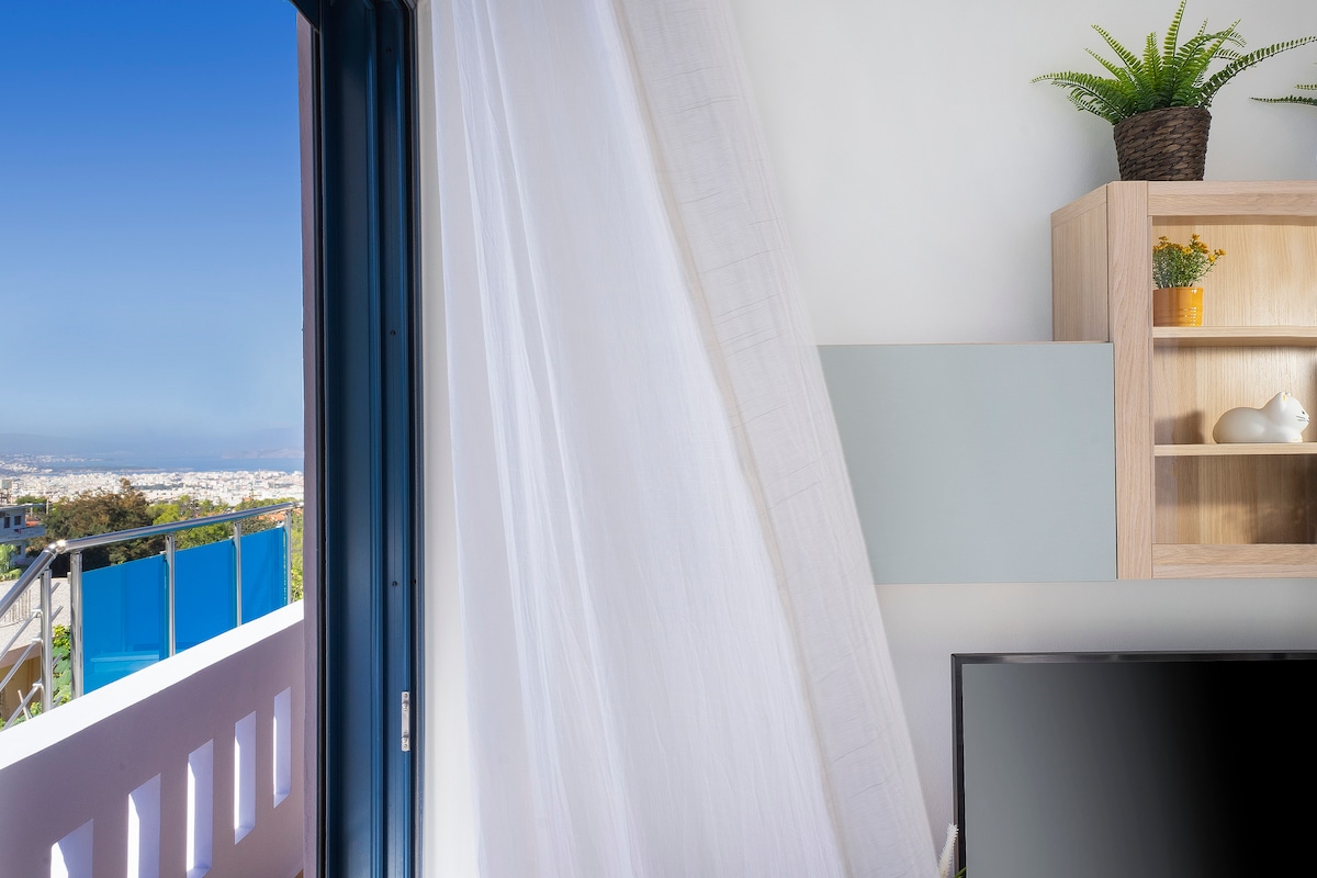 A light curtain partially obscures a modern living space, revealing a stunning view of the city and mountains through an open door. A wooden shelf with decorative items stands against a neutral wall, contributing to the airy ambiance.