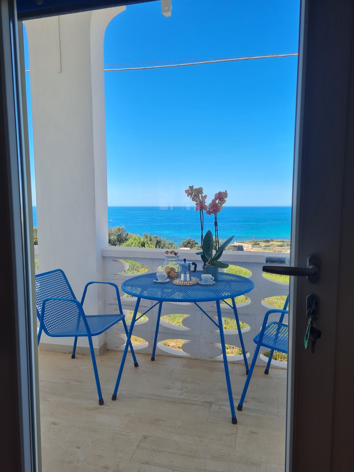 A small terrace is visible through an open door, featuring a blue table surrounded by two matching chairs. A flower arrangement sits atop the table, complemented by decorative items. Beyond, a panoramic view of the sea stretches under a clear blue sky.