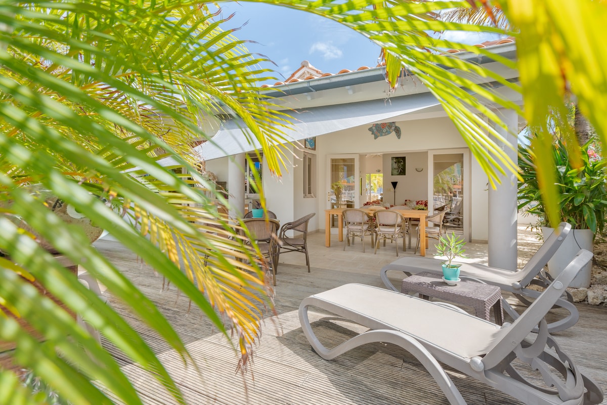 A spacious outdoor terrace is framed by tropical plants, offering a view of a cozy seating area with a dining table. Sun loungers are positioned nearby, inviting relaxation. The structure features an open design, enhancing the connection between indoor and outdoor spaces.