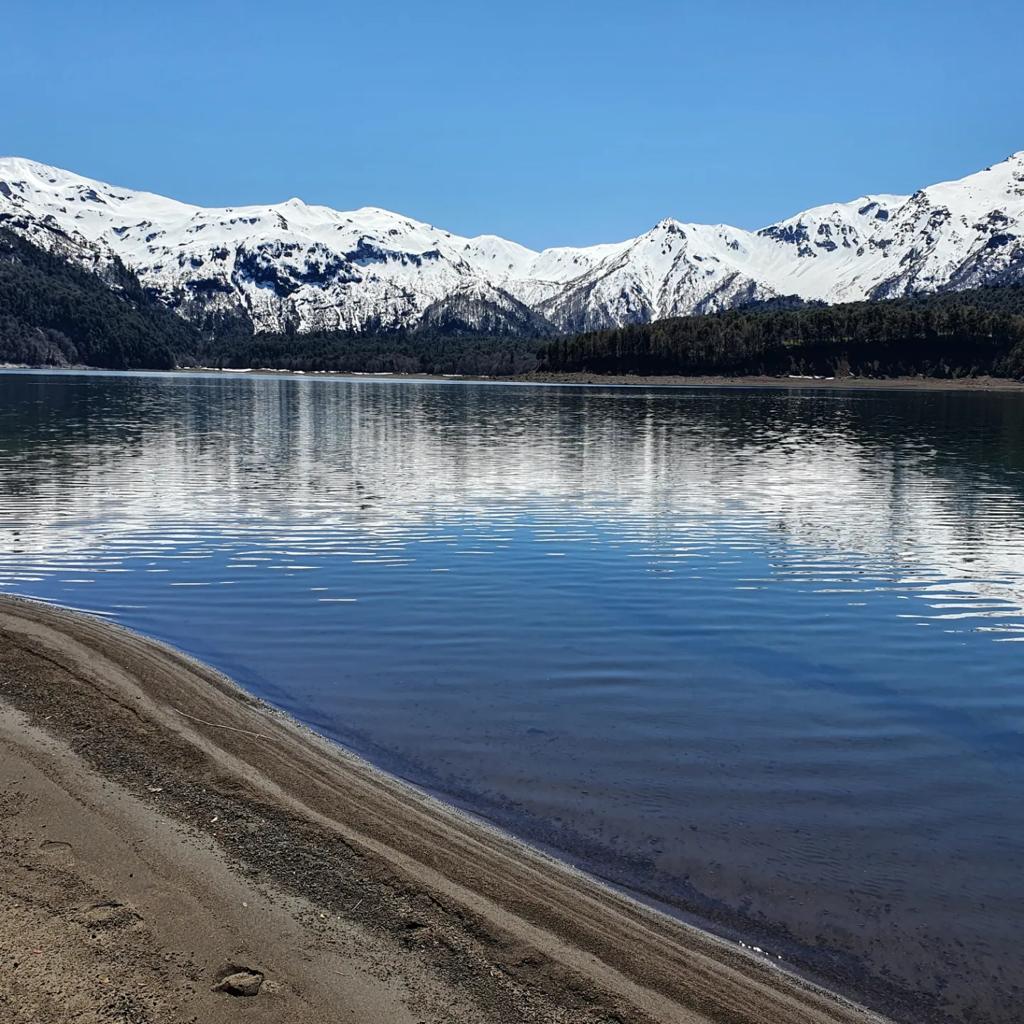 A serene lake reflects a clear blue sky and snow-capped mountains in the background. The water surface displays gentle ripples, while a sandy shoreline curves along the edge, providing a natural border to the tranquil landscape.