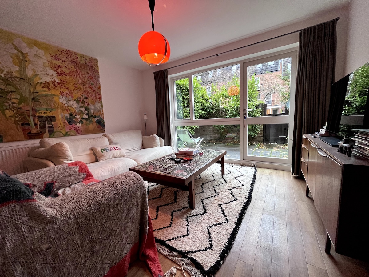 A welcoming living area features a light-colored sofa and a decorative rug on wooden flooring. Large glass doors open to a private garden, allowing natural light to fill the space. A vibrant artwork adorns the wall, adding a touch of color to the room.