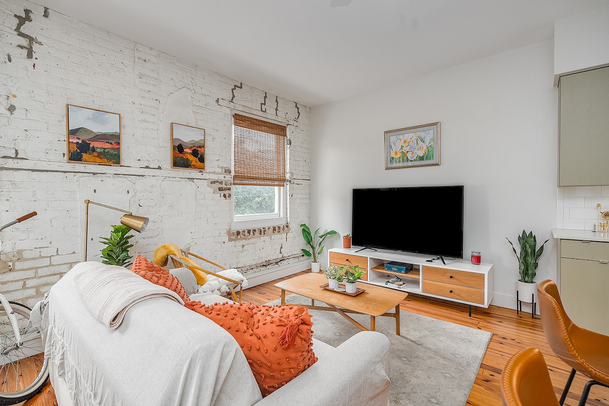 The living area features exposed brick walls complemented by a light, neutral color palette. A comfortable sofa adorned with colorful throw pillows sits atop a soft area rug. A flat-screen TV is mounted on a wooden media console, with greenery and artwork adding warmth to the space.