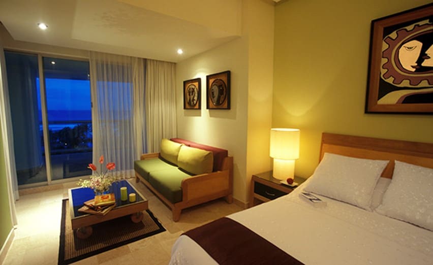 A spacious room features a king-size bed with crisp linens, complemented by a cozy seating area with a green sofa. Two framed artworks adorn the walls, and a table is set with decorative items. A large window reveals an evening view of the beach.
