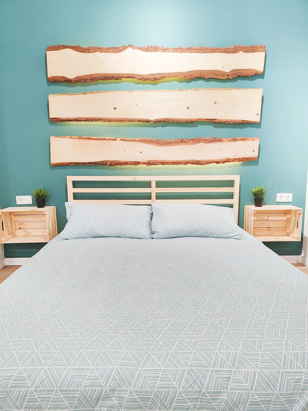 A neatly arranged sleeping area features a spacious bed with a light blue patterned coverlet. Natural wood accents are displayed on the wall above the headboard. Small wooden nightstands on either side hold potted plants, contributing to a calm and inviting environment.