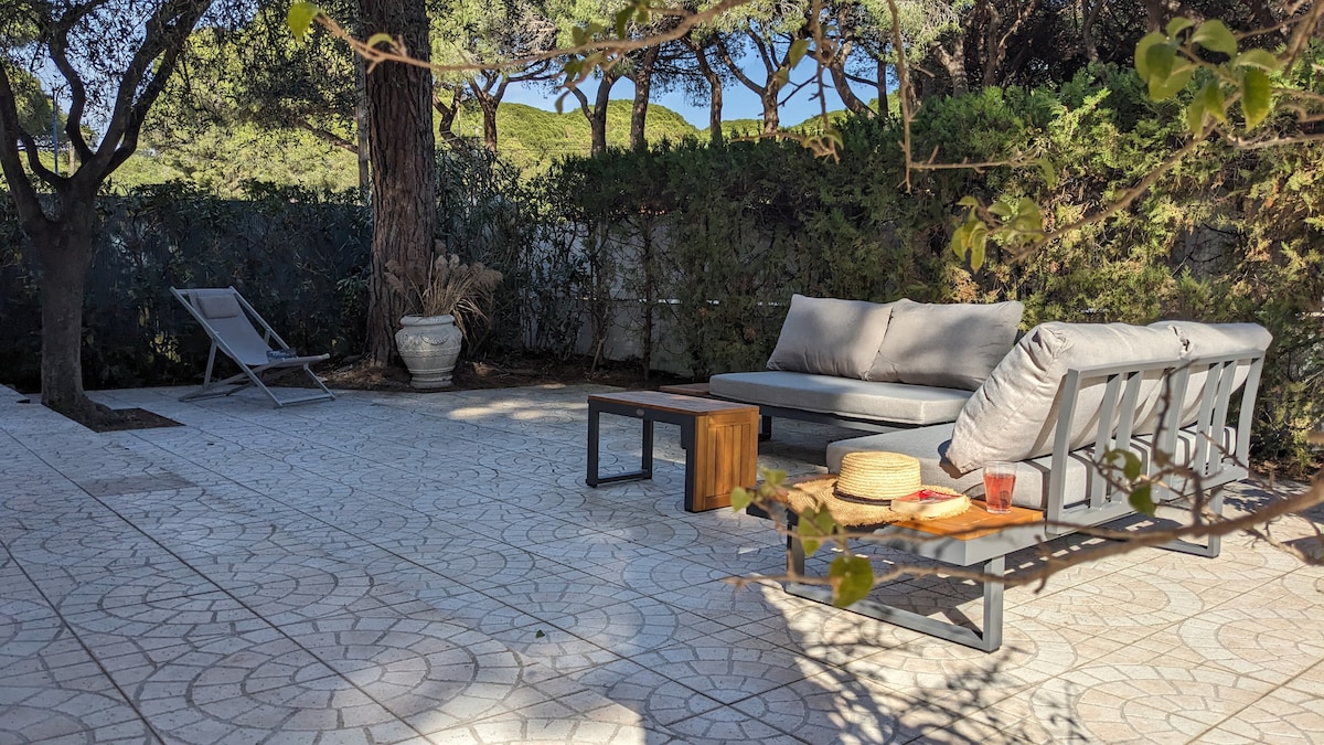 A serene outdoor space is presented, featuring a seating area with a comfortable sofa and a wooden coffee table. A sun lounger is positioned nearby on the patterned tile flooring. Lush greenery surrounds the area, providing privacy and shade.