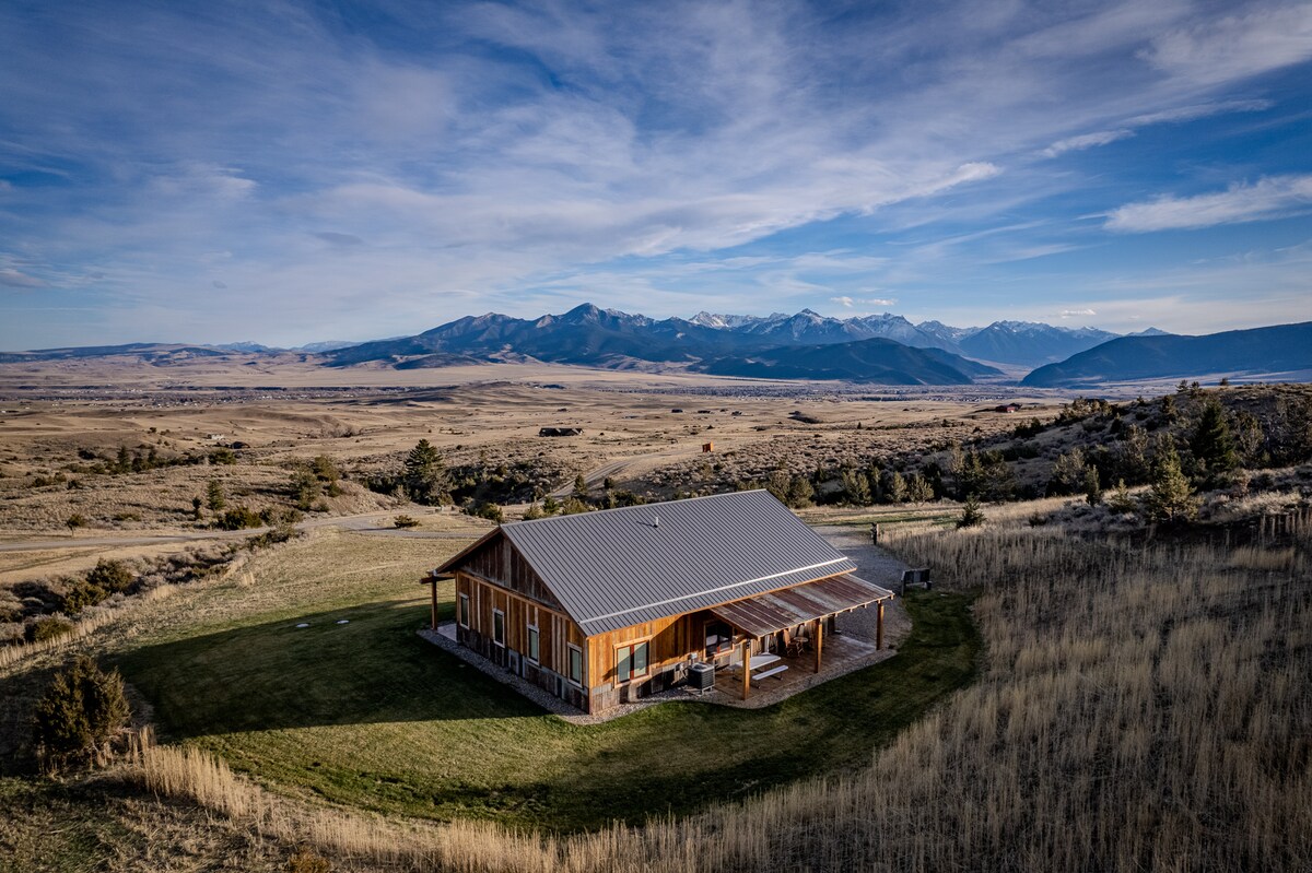 A cabin with a timber frame design is set against a backdrop of distant mountains and open fields. The expansive rural landscape is dotted with vegetation, showcasing the natural beauty of the surroundings. A clear sky enhances the serenity of this private retreat.