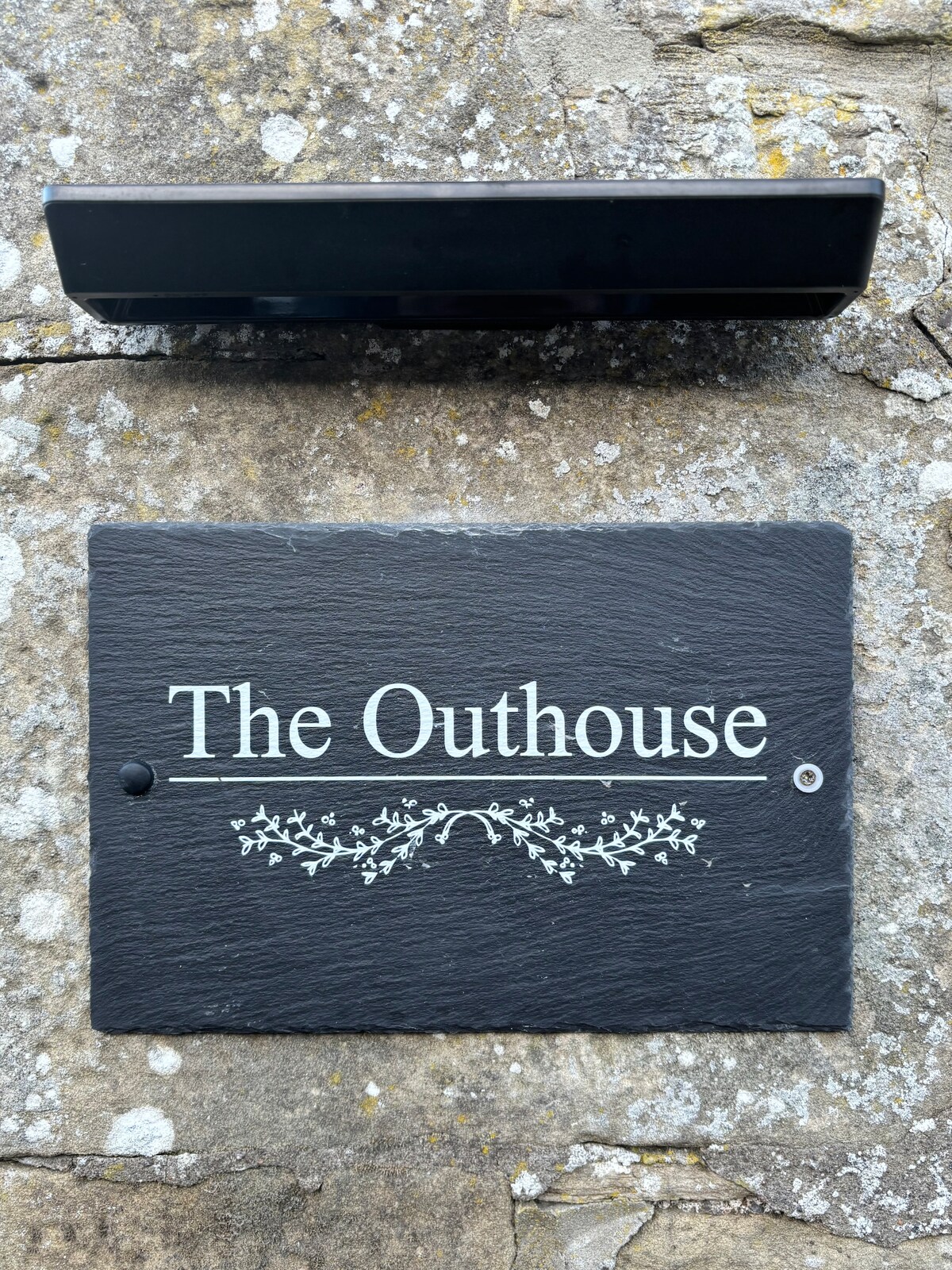 A slate sign reading 'The Outhouse' is mounted against a textured stone wall. The sign features decorative white floral embellishments, providing a clear indication of the cottage's identity.