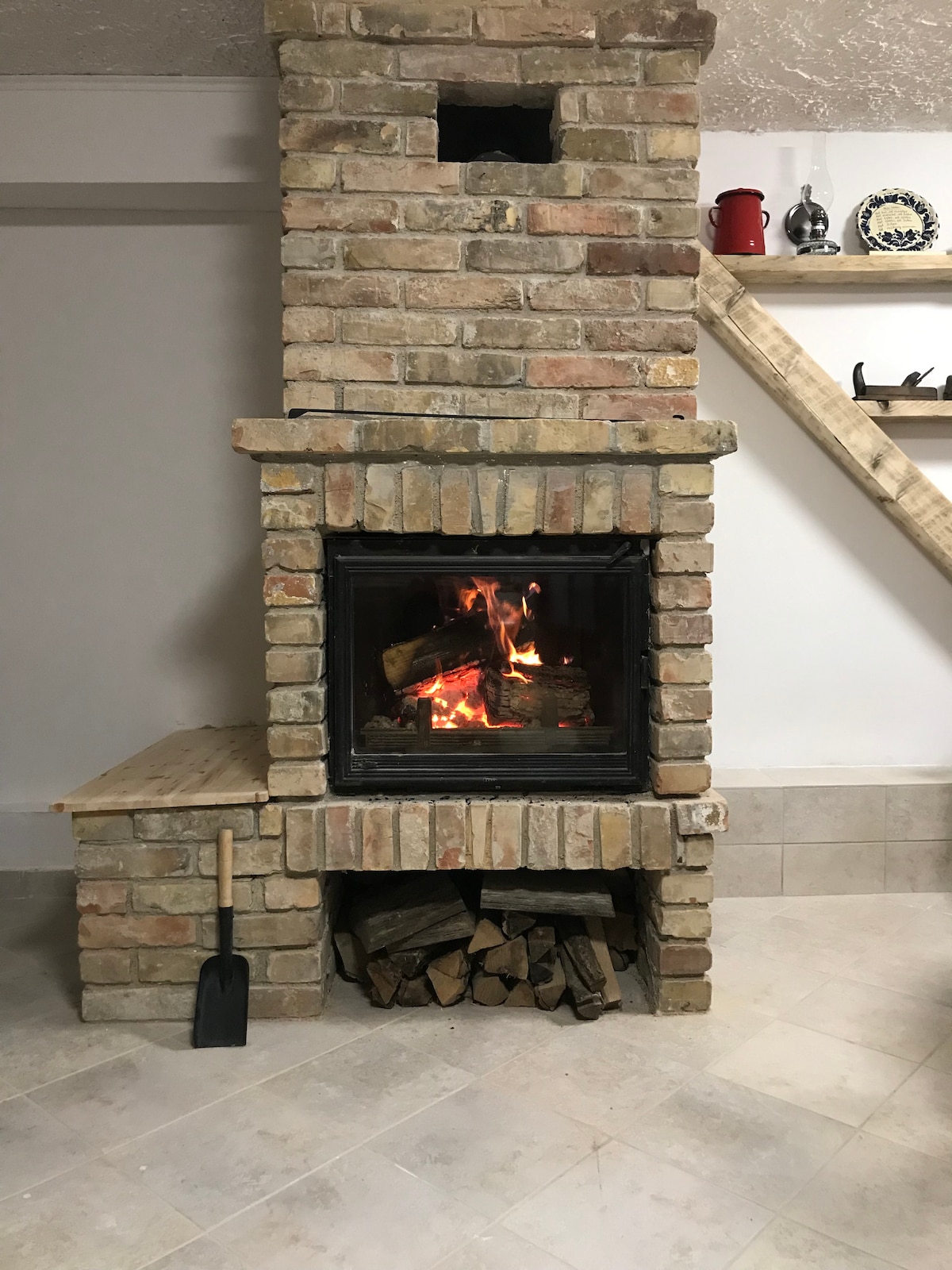 A rustic brick fireplace is centered in the image, with flames visible within the glass enclosure. A wooden shelf above displays various items, while a stack of firewood is stored beneath the fireplace. A metal shovel rests on the floor beside the firewood.